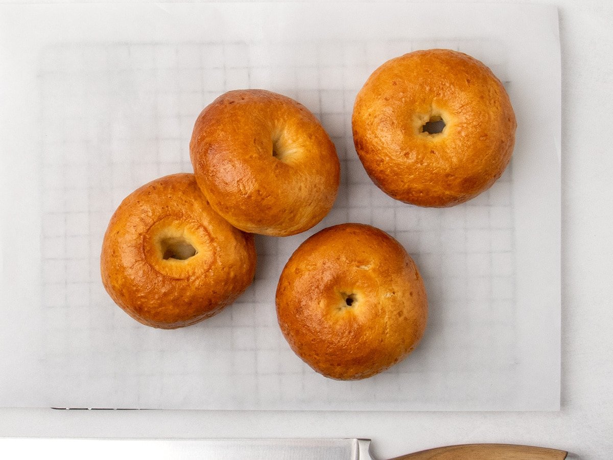 Four golden brown gluten free bagels on white parchment paper with the edge of a large bread knife visible at the bottom.