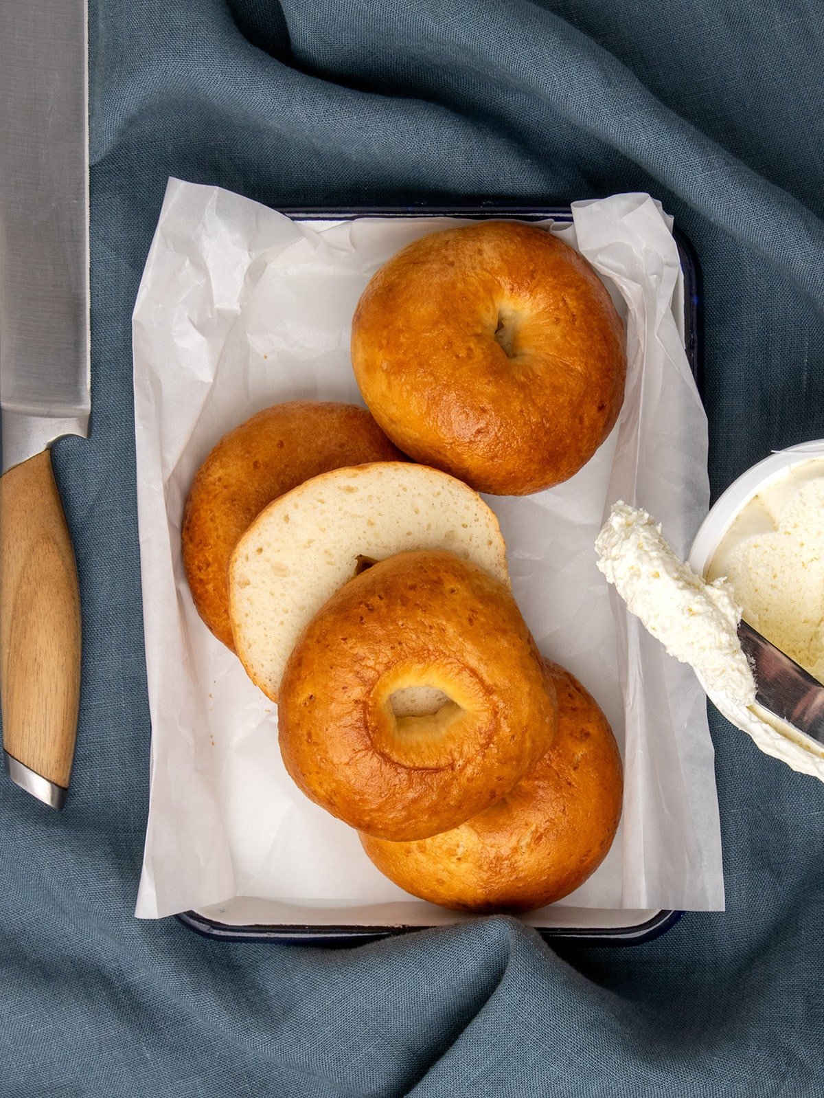 Three whole golden brown bagels in a parchment-lined small white baking sheet, with one sliced bagel showing the inside and a knife with a spread of white cream beside it.