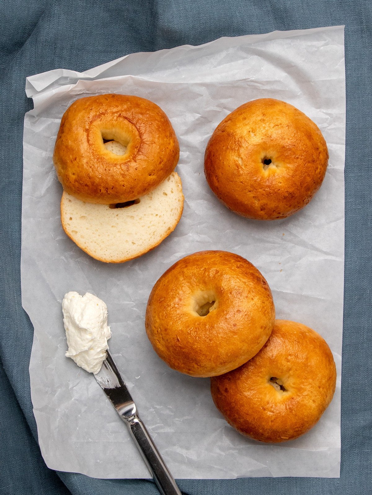 Four golden brown bagels, one sliced in half to reveal a chewy inside crumb, on white parchment paper with a knife holding a dollop of white cream nearby.