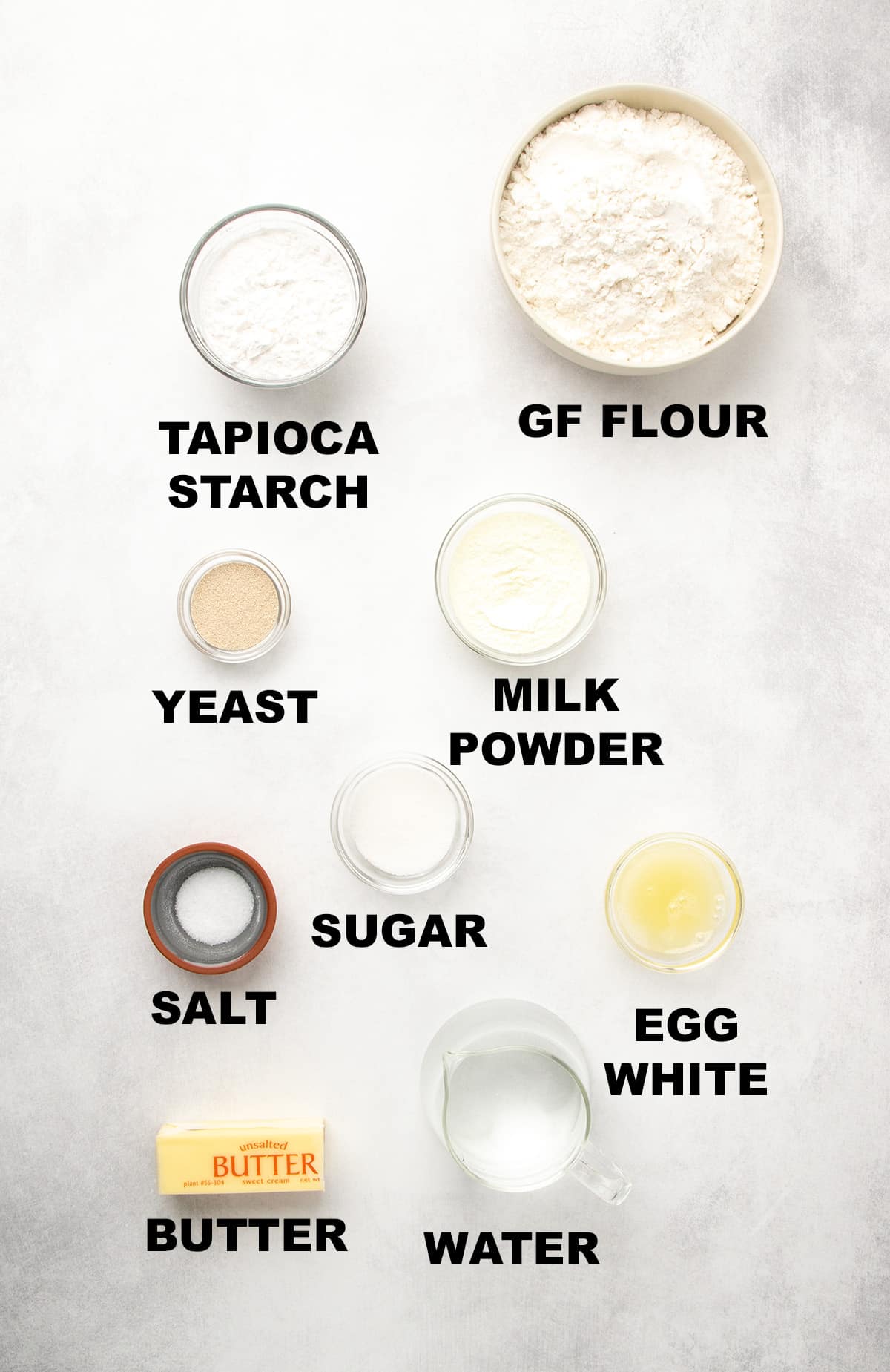 Bowls and small dishes of dry and wet ingredients arranged on a white surface, each labeled with text including “TAPIOCA STARCH,” “GF FLOUR,” “YEAST,” “MILK POWDER,” “SALT,” “SUGAR,” “BUTTER,” “WATER,” and “EGG WHITE.”