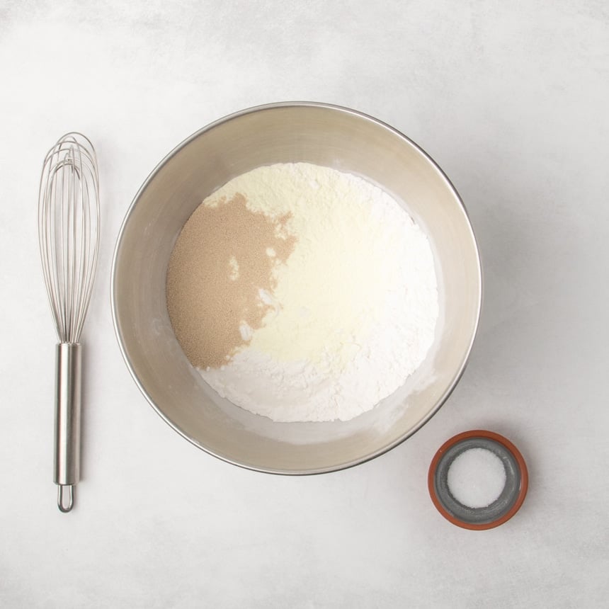 Metal mixing bowl filled with white flour and a mound of instant yeast granules, with a whisk beside the bowl and a small dish of salt on a light surface.