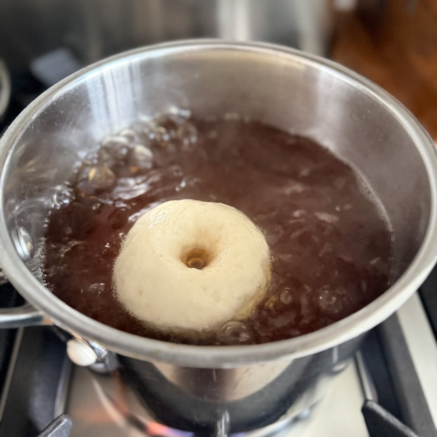 Ring-shaped swollen dough piece floating in a pot of dark boiling liquid on a stovetop.