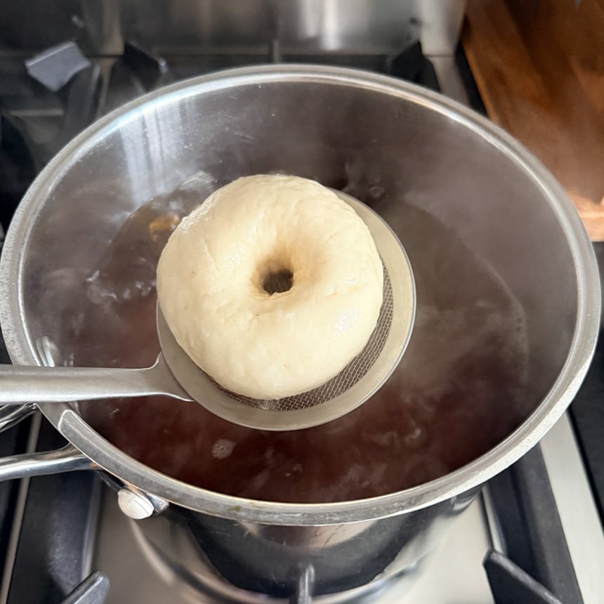 Swollen ring-shaped dough piece lifted on a mesh skimmer above a pot of dark boiling liquid.