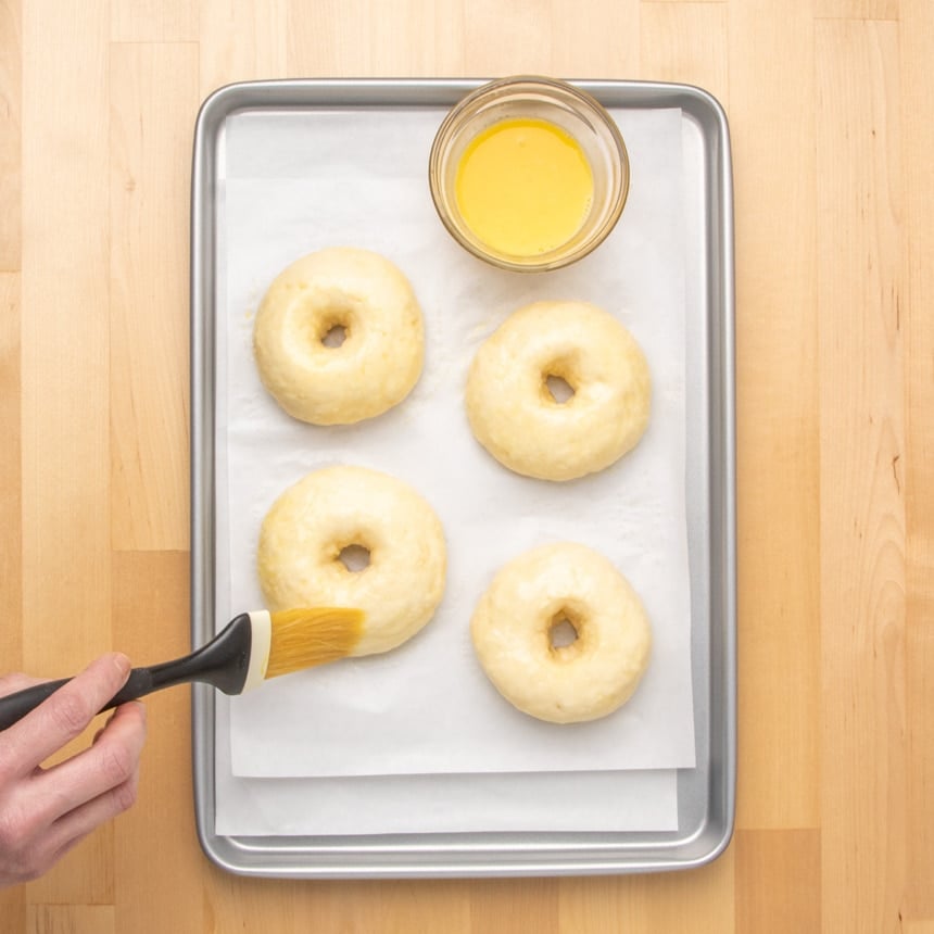 Four ring-shaped risen raw dough pieces on a parchment-lined baking sheet as a hand brushes one with a pastry brush, with a small bowl of yellow liquid egg wash nearby.