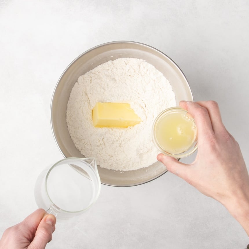 Metal mixing bowl with dry ingredients and a stick of butter inside, as one hand pours water from a measuring cup and another hand holds a small glass bowl with an egg white above the bowl.