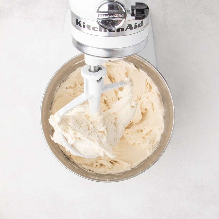 Stand mixer with paddle attachment after mixing thick, pale raw dough in a metal mixing bowl that's sticking to the paddle and the sides of the mixing bowl, looking slightly whipped and aerated.