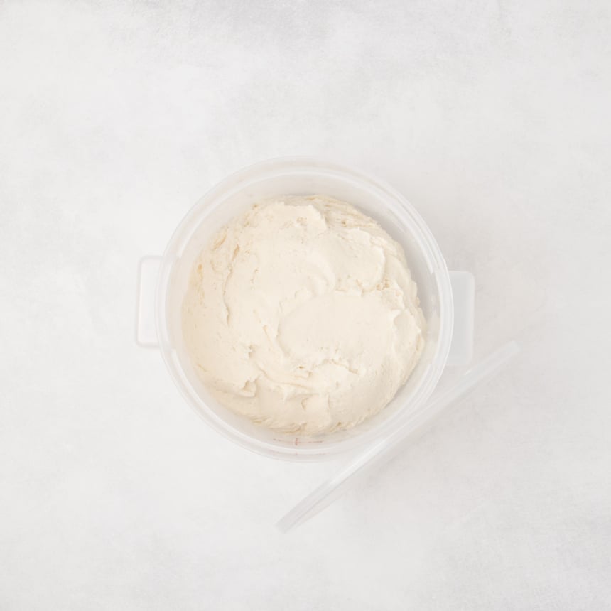 Round plastic container filled with raw smooth pale dough and a lid placed beside it on a light surface.