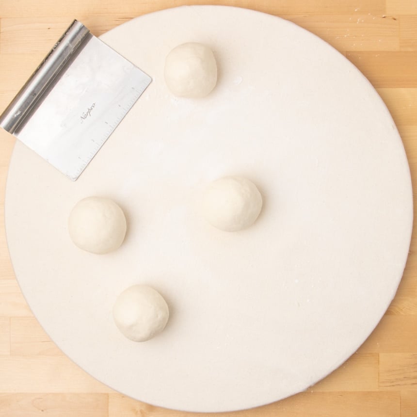 Round white board with four smooth dough balls spaced apart and a metal bench scraper resting nearby.