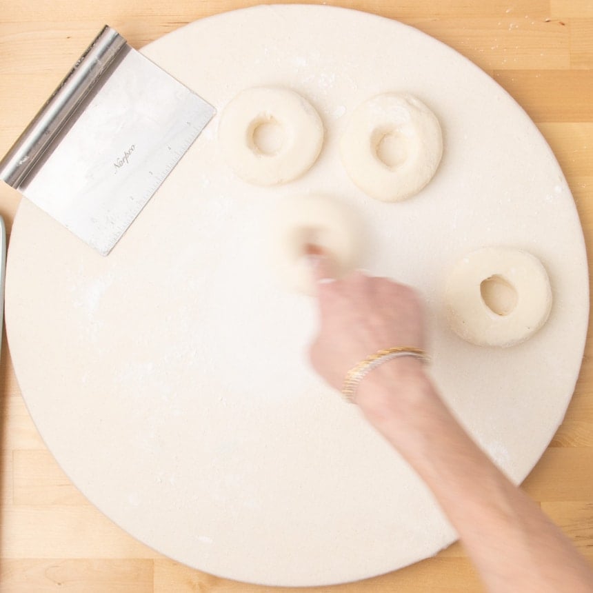 Round white board with three dough rounds shaped into round disks with holes in the center, as the index finger of a hand rotates around in the center of the 4th piece of raw dough.