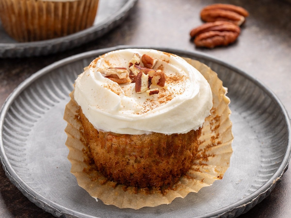 Close-up of a frosted carrot cake cupcake in a paper liner on a metal plate, topped with chopped nuts and a dusting of ground cinnamon.