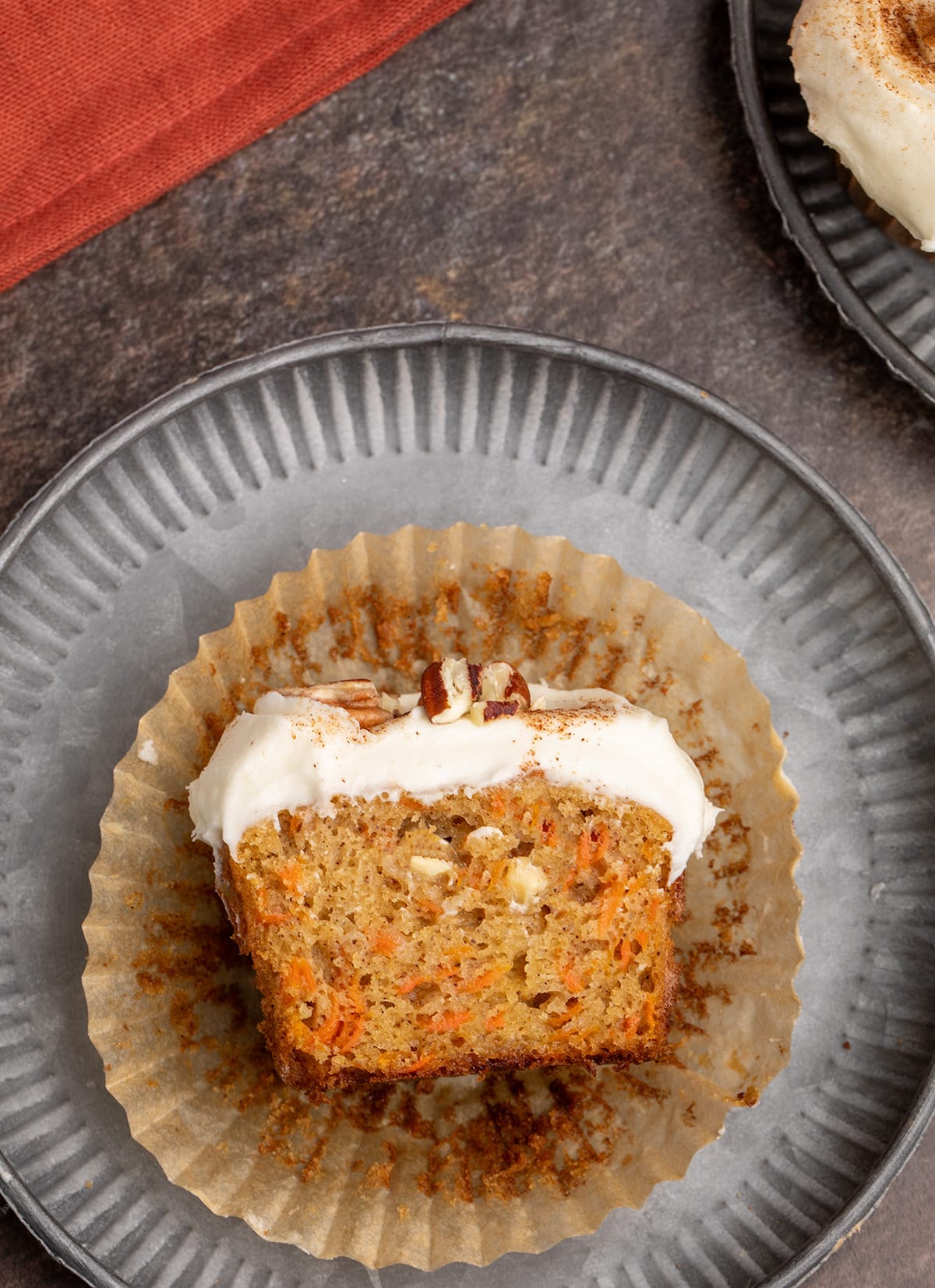 Overhead view of a carrot cupcake cut in half in its paper liner on a metal plate, showing the interior with shredded carrots and white chips.