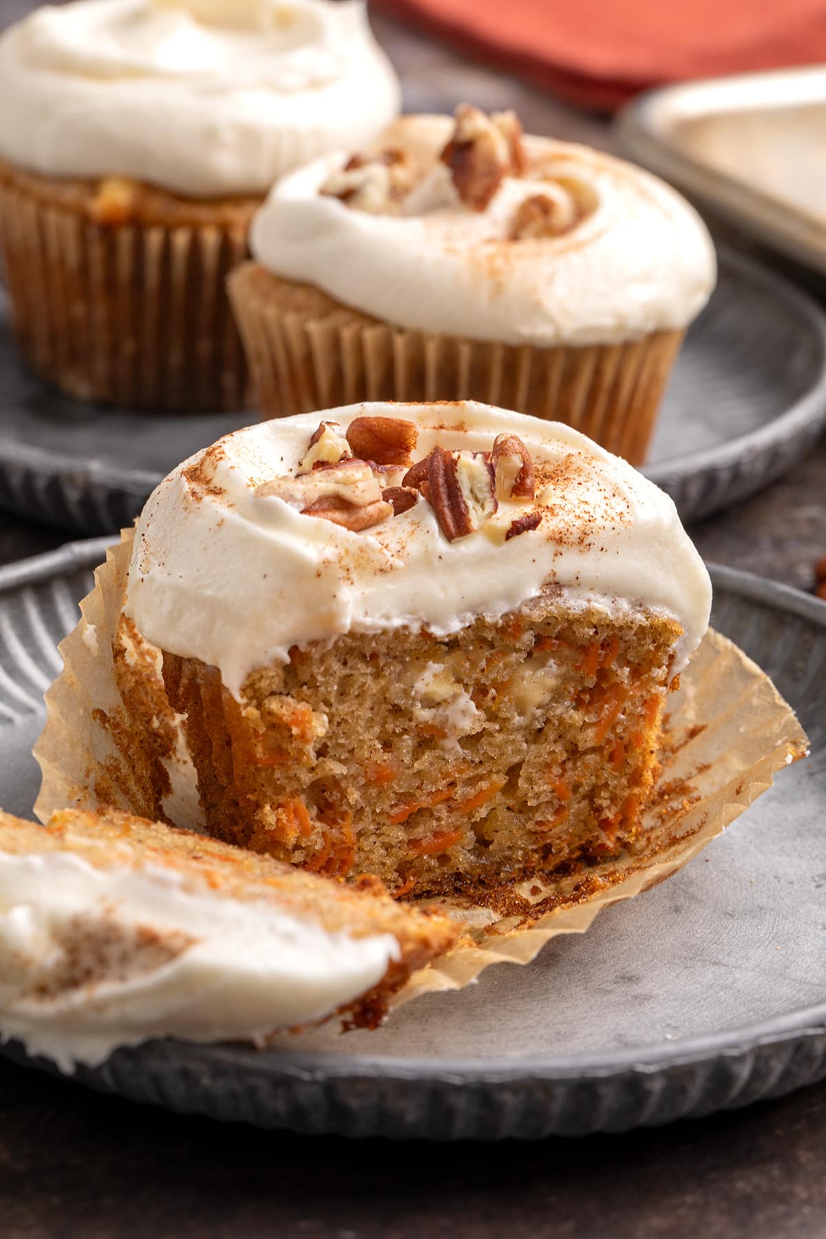Close-up of a cut carrot cake cupcake with cream-colored frosting and chopped nuts on top, with additional frosted cupcakes in the background.