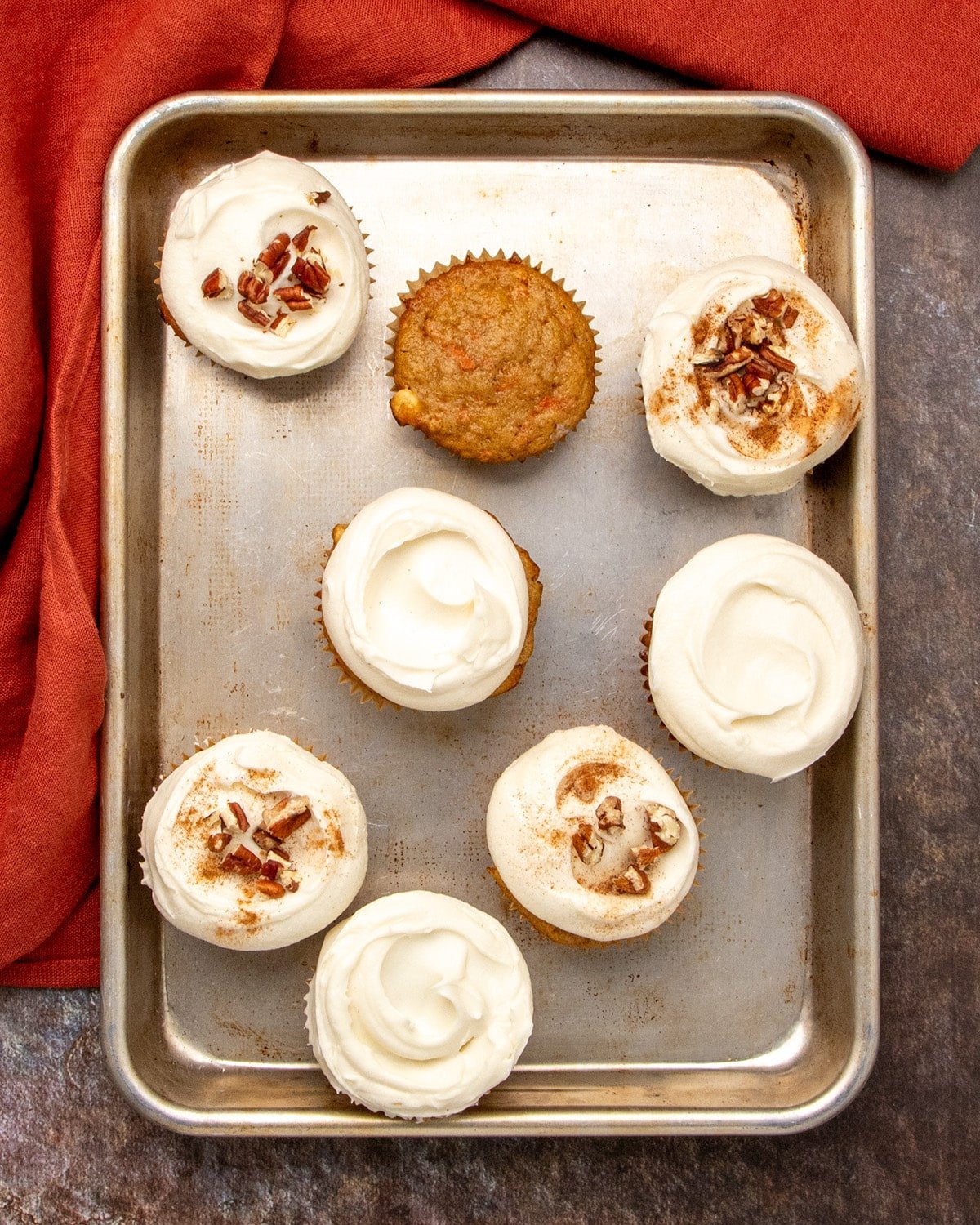 Overhead view of a metal baking sheet holding 7 frosted carrot cake cupcakes and one plain, with an orange-red cloth beside the pan.
