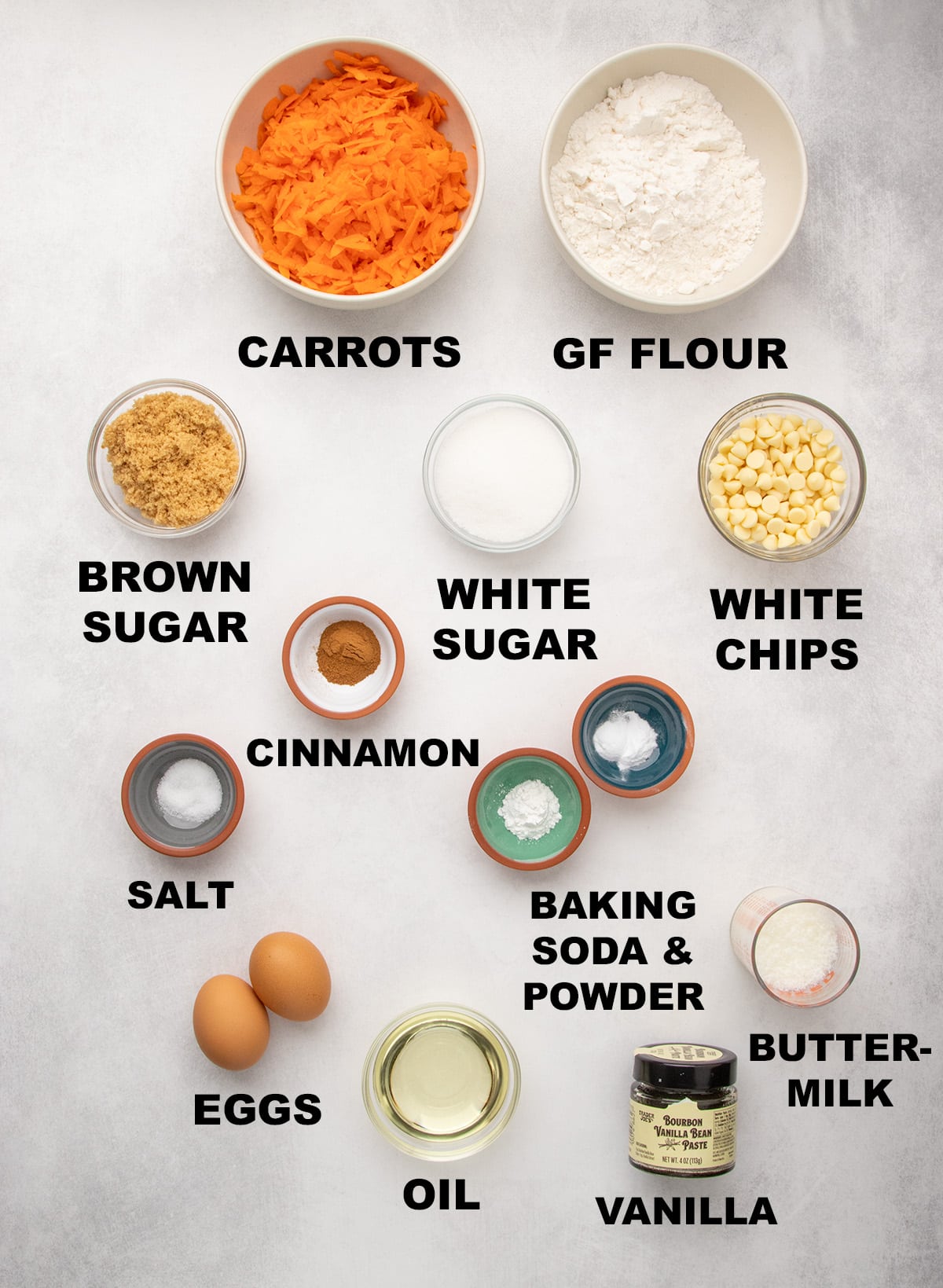 Overhead view of small bowls and other containers of shredded carrots, gluten free flour, brown sugar, white sugar, white chips, cinnamon, salt, baking soda and baking powder, eggs, oil, buttermilk, and vanilla, each labeled with text.