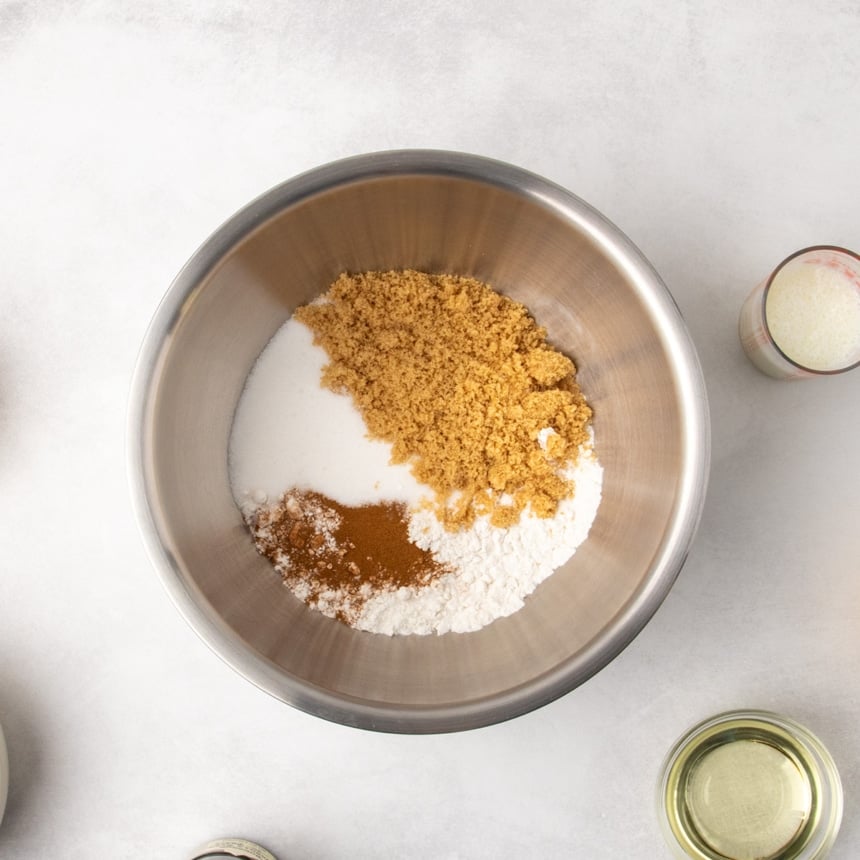 Overhead view of a metal mixing bowl filled with white sugar, brown sugar, white flour, and a ground cinnamon, with a glass measuring cup of buttermilk and a small bowl of oil nearby on a light surface.