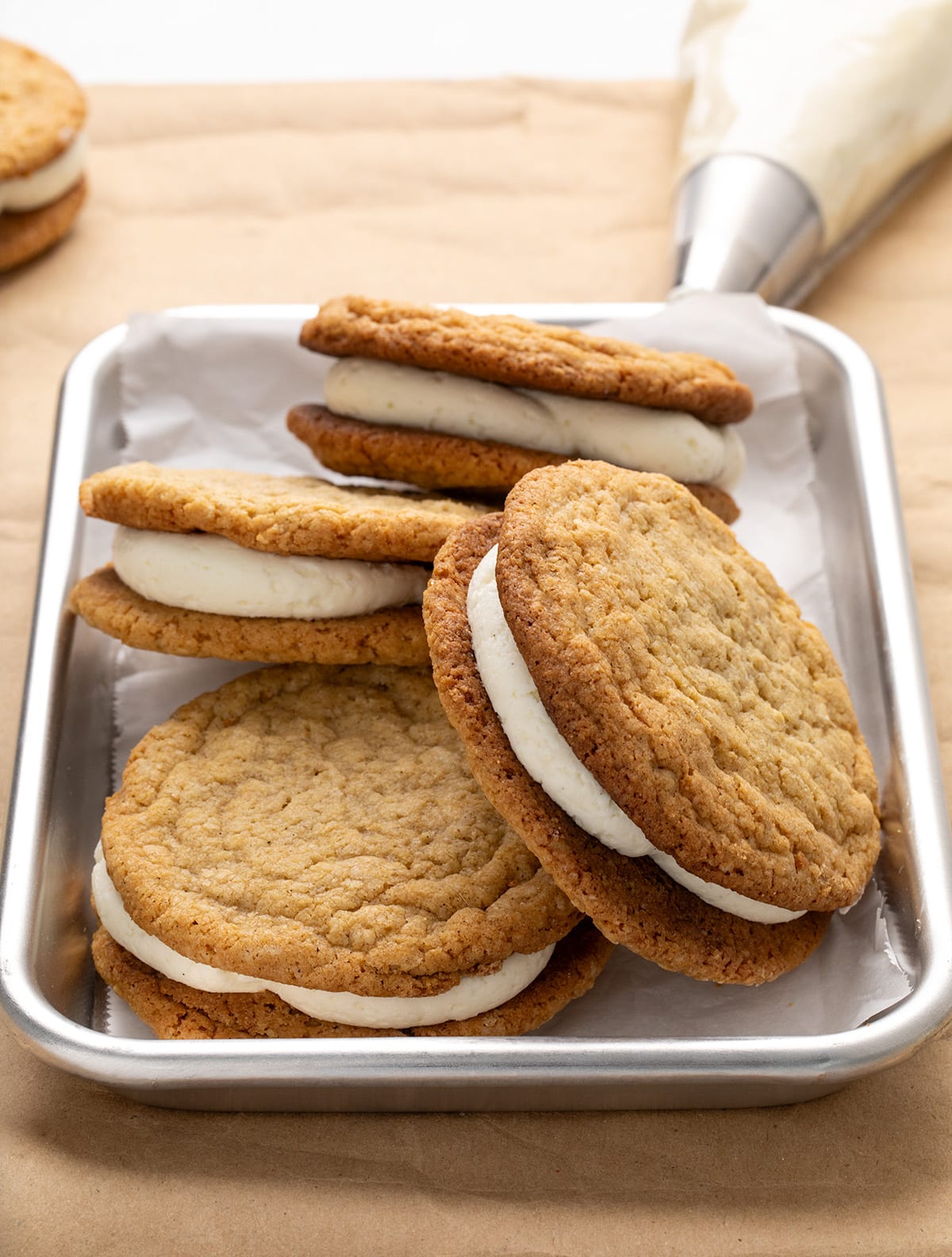 Several brown oatmeal cream pie sandwiches overlapping on a small white parchment paper-lined metal tray on brown kraft paper, with a piping bag in the background, viewed from the side.