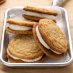 Close-up of stacked oatmeal cream pie sandwiches on parchment paper, showing thick white cream filling between golden brown cookies, with a piping bag blurred in the background.