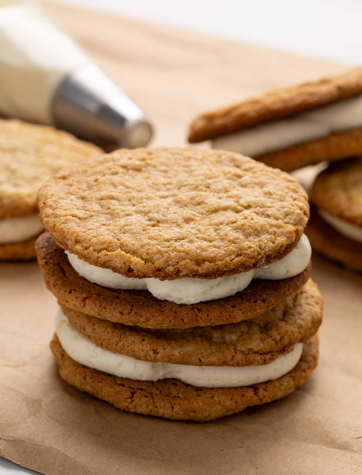 Two oatmeal cream pie sandwiches stacked vertically on brown kraft paper, with additional sandwiches and a piping bag in the background.