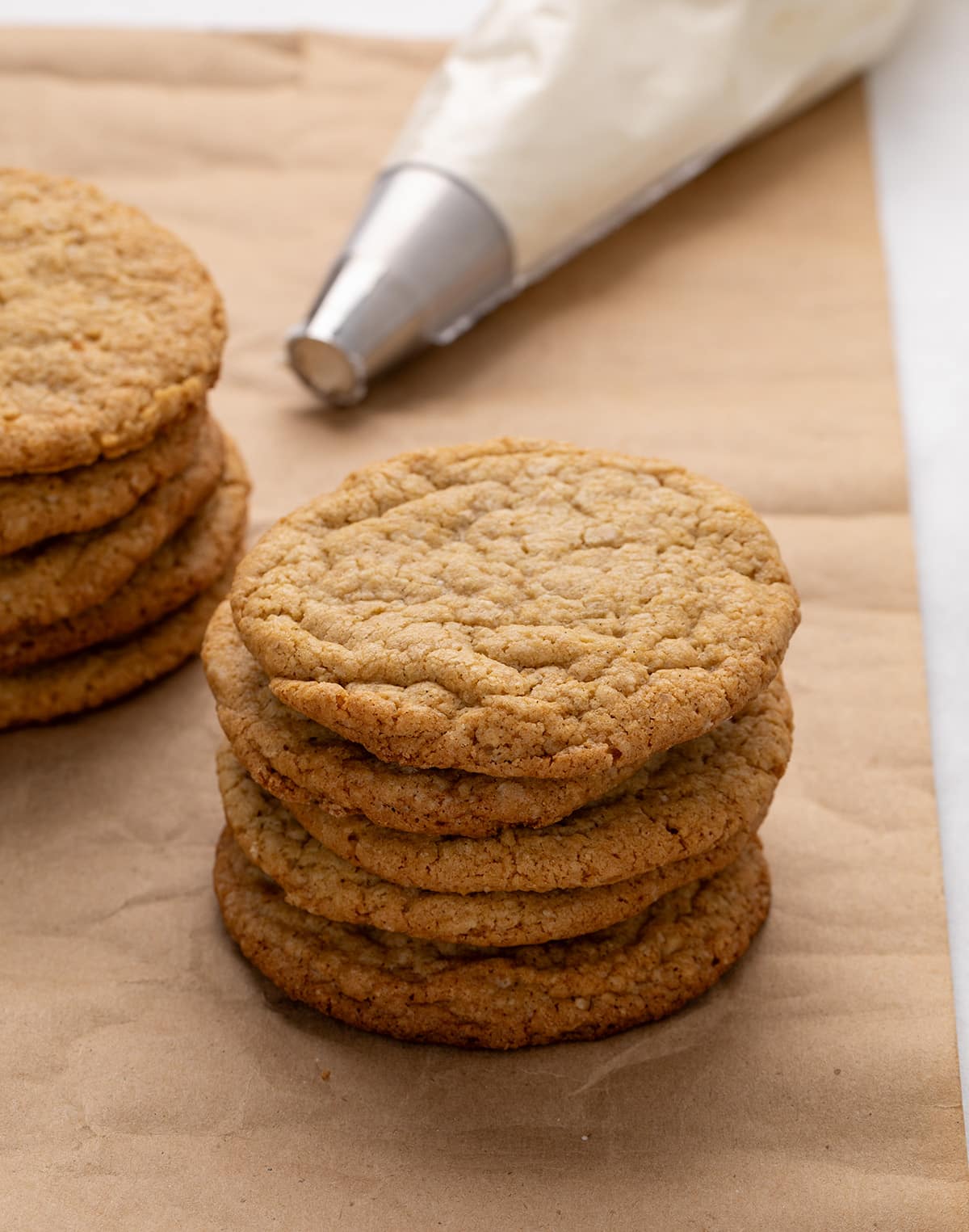 Vertical stack of 5 plain golden brown baked round cookies on brown kraft paper with another stack of plain cookies beside it and a piping bag in the background.