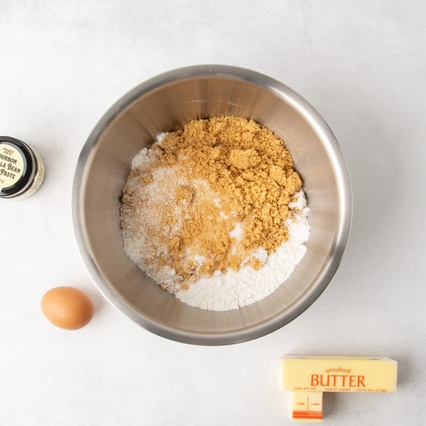 Overhead view of a metal mixing bowl filled with white powder ingredients, ground oats, brown sugar, with a whole egg, a wrapped stick of butter labeled unsalted, and a small jar of vanilla bean paste beside the bowl.