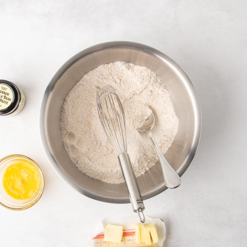 Overhead view of a metal mixing bowl with dry ingredients mixed together, a whisk and spoon resting inside, a small bowl of beaten egg, sliced butter on its wrapper, and a jar of vanilla bean paste nearby.