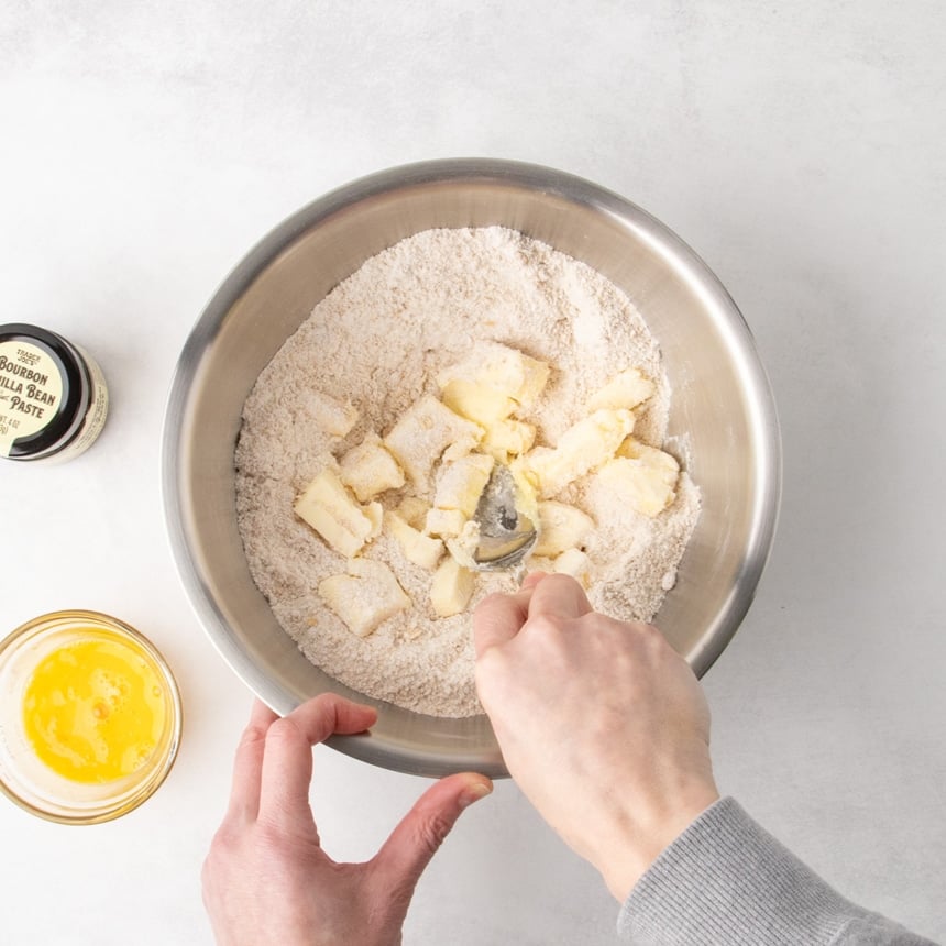 Overhead view of a hand using a spoon to press chunks of butter into dry ingredients in a metal mixing bowl.