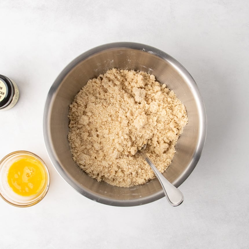 Overhead view of a metal mixing bowl filled with a very light brown crumbly mixture and a spoon resting inside.