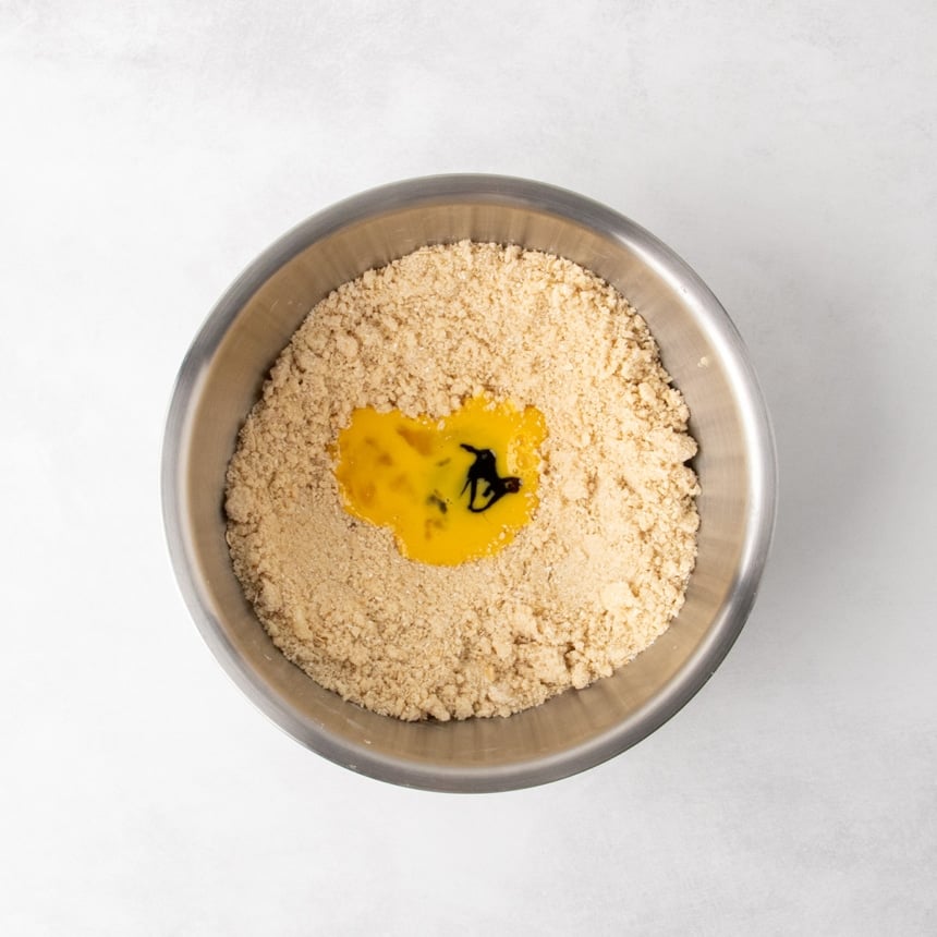 Overhead view of a metal mixing bowl with a well in the crumbly mixture holding beaten egg and a splash of dark vanilla bean paste in the center.