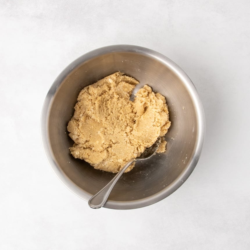 Overhead view of a metal mixing bowl containing a thick, cohesive raw cookie dough with a spoon resting inside.