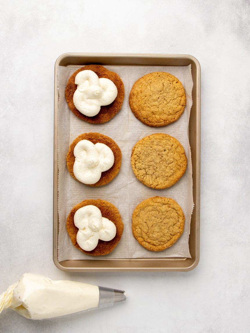 Overhead view of a parchment-lined baking sheet with six cookies, three turned over and topped with swirls of white frosting and three plain, with a plastic piping bag of frosting nearby.