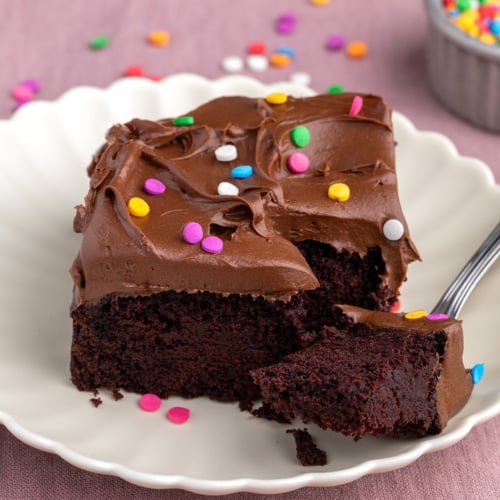 Closeup image of a square slice of chocolate cake with chocolate frosting and colorful sprinkles on a white plate, with a piece on a fork that is set in front.