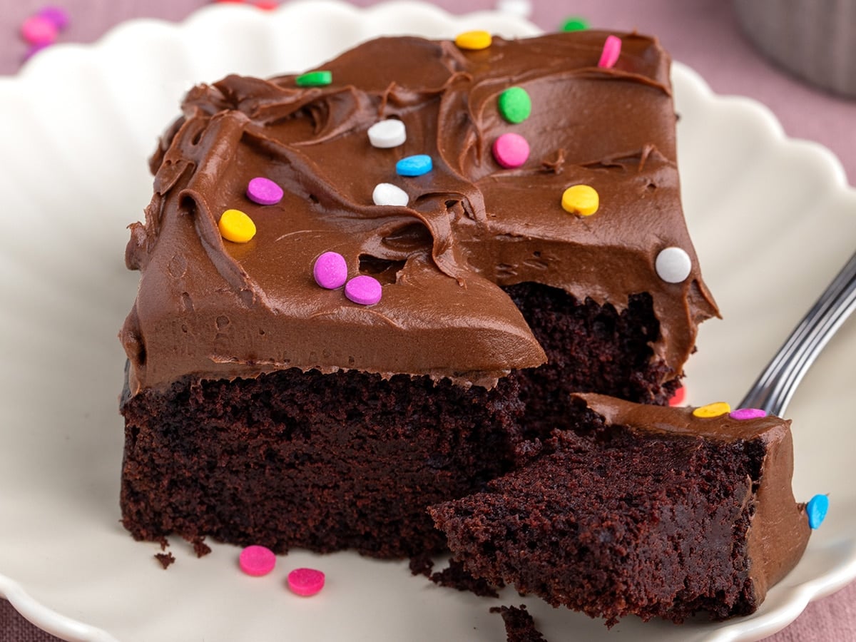 A square slice of gluten free dairy free chocolate cake with chocolate frosting and colorful sprinkles on a white plate with a small bite taken still on a fork, with sprinkles scattered in the background.
