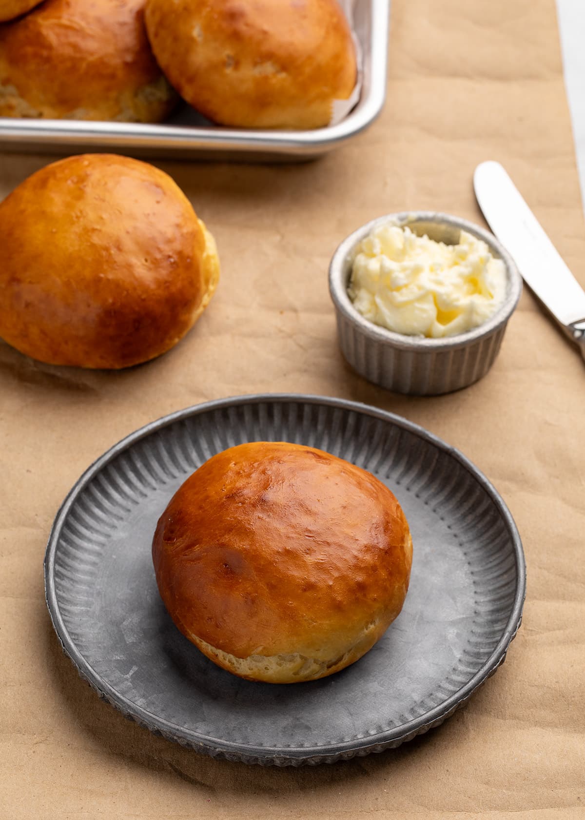 Single golden brown roll on a gray plate with additional rolls, a small dish of butter, and a butter knife on a brown kraft paper surface.
