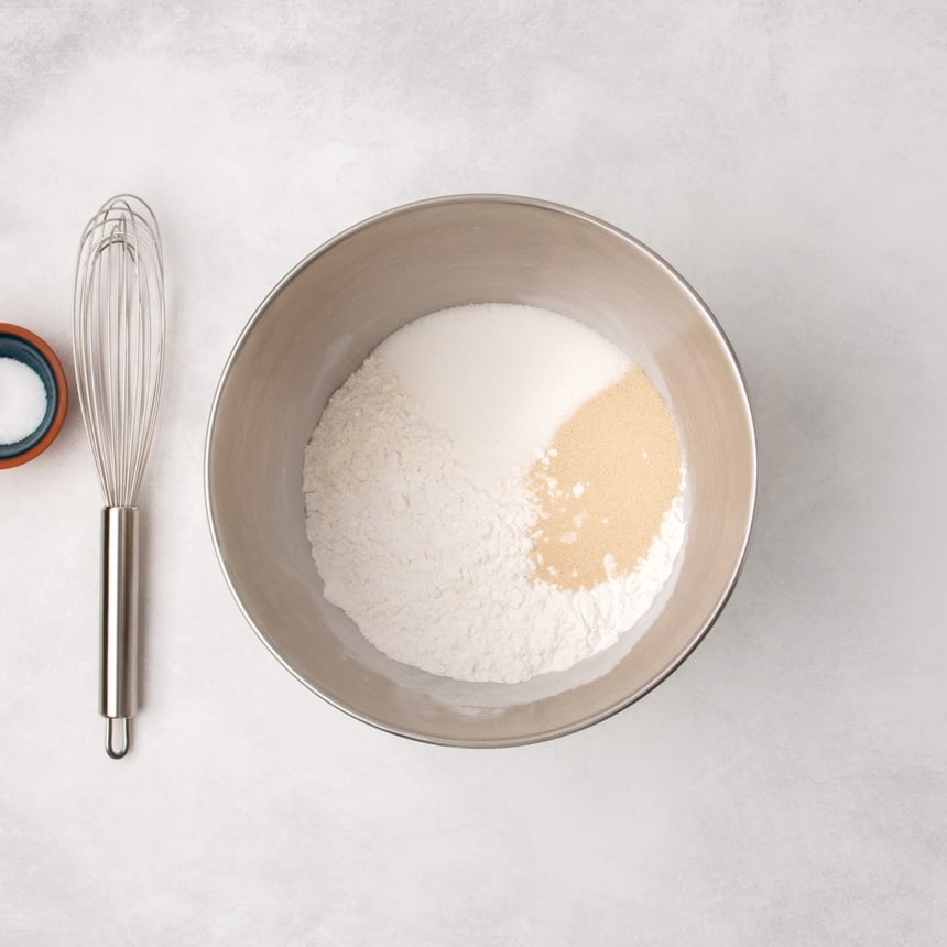 Metal mixing bowl with unwhisked white flour, granulated sugar, and a instant yeast, with a whisk and a small bowl of salt beside it.