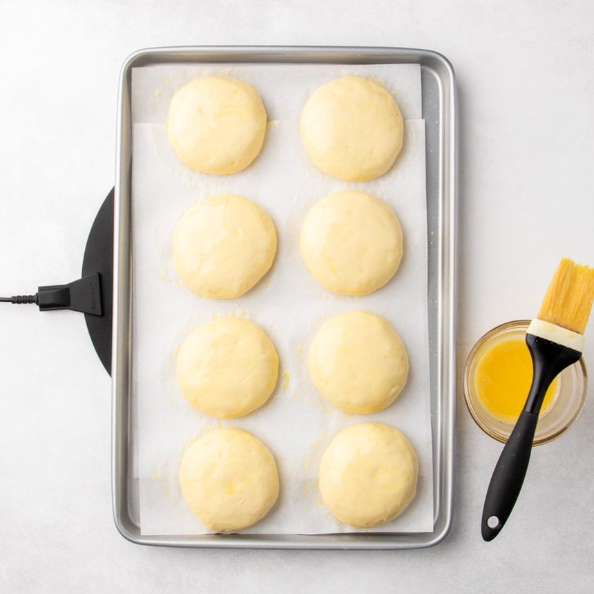 Shaped dough rounds on a parchment-lined baking sheet brushed with egg wash, with a bowl of the remaining egg wash and a pastry brush beside it.