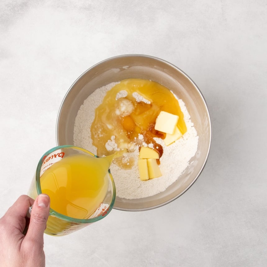 Wet ingredients including eggs, butter, and vanilla added to the dry mixture while a hand pours orange-yellow liquid from a measuring cup into the bowl.