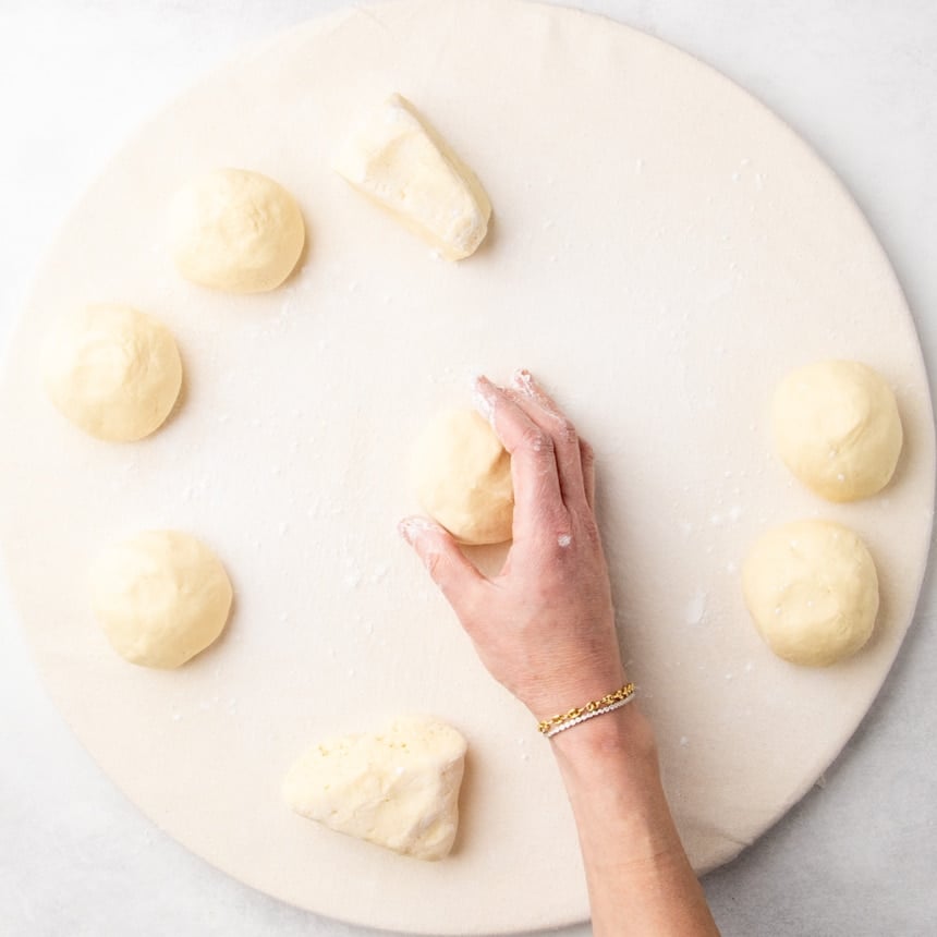 Several small dough portions shaped into balls on a floured cloth surface, with a hand shaping one piece.
