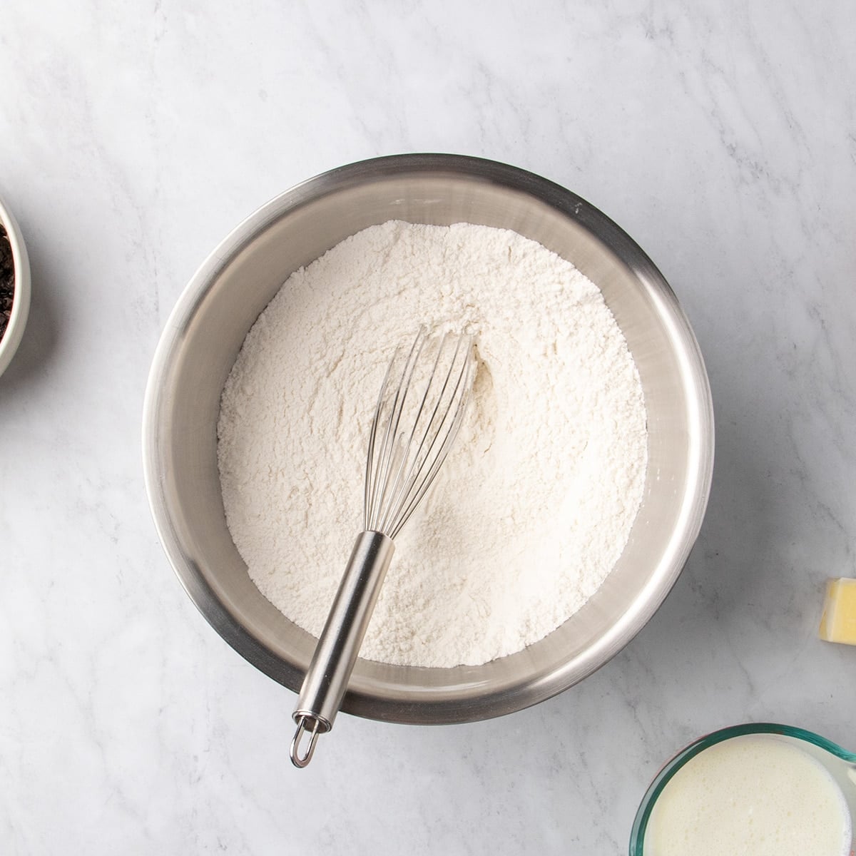 Metal mixing bowl filled with white dry ingredients and a metal whisk resting in the center on a marble surface.