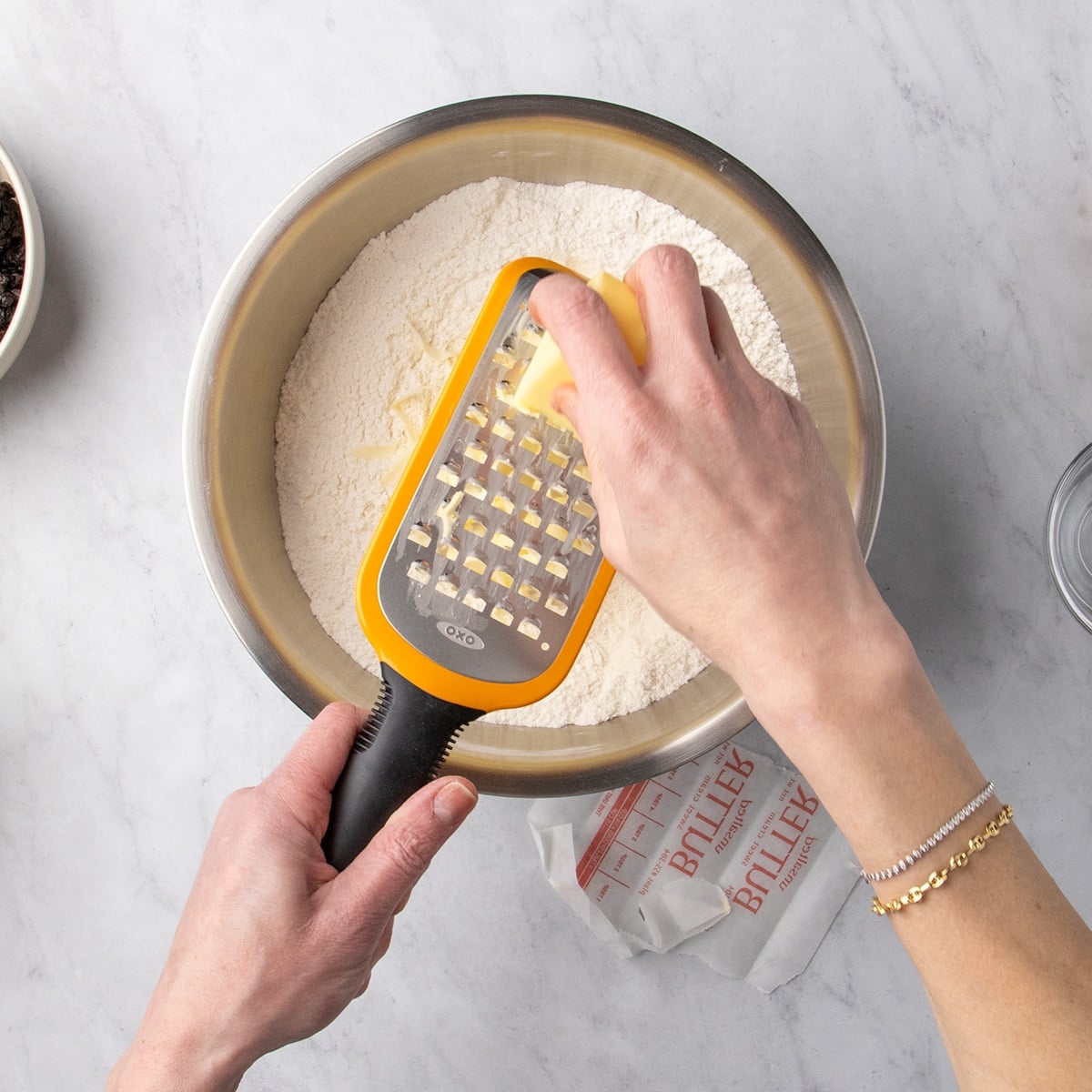 Hands grating a stick of butter on a coarse grater directly into a bowl of white dry ingredients.