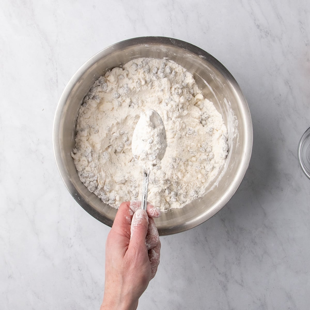 Hand holding a spoon over a bowl of dry ingredients mixed with grated butter and raisins with small coated pieces visible.