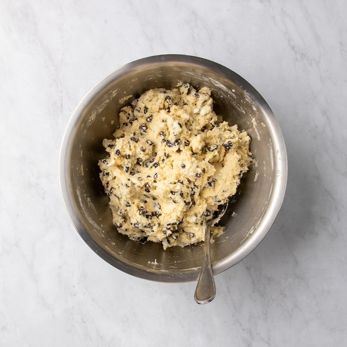 Thick raw soda bread dough with small dark pieces of raisins mixed throughout in a metal bowl with a spoon.