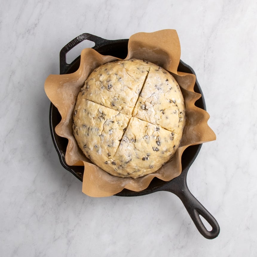 Round of dough with a cross cut into the top placed in a parchment-lined black round cast iron skillet.