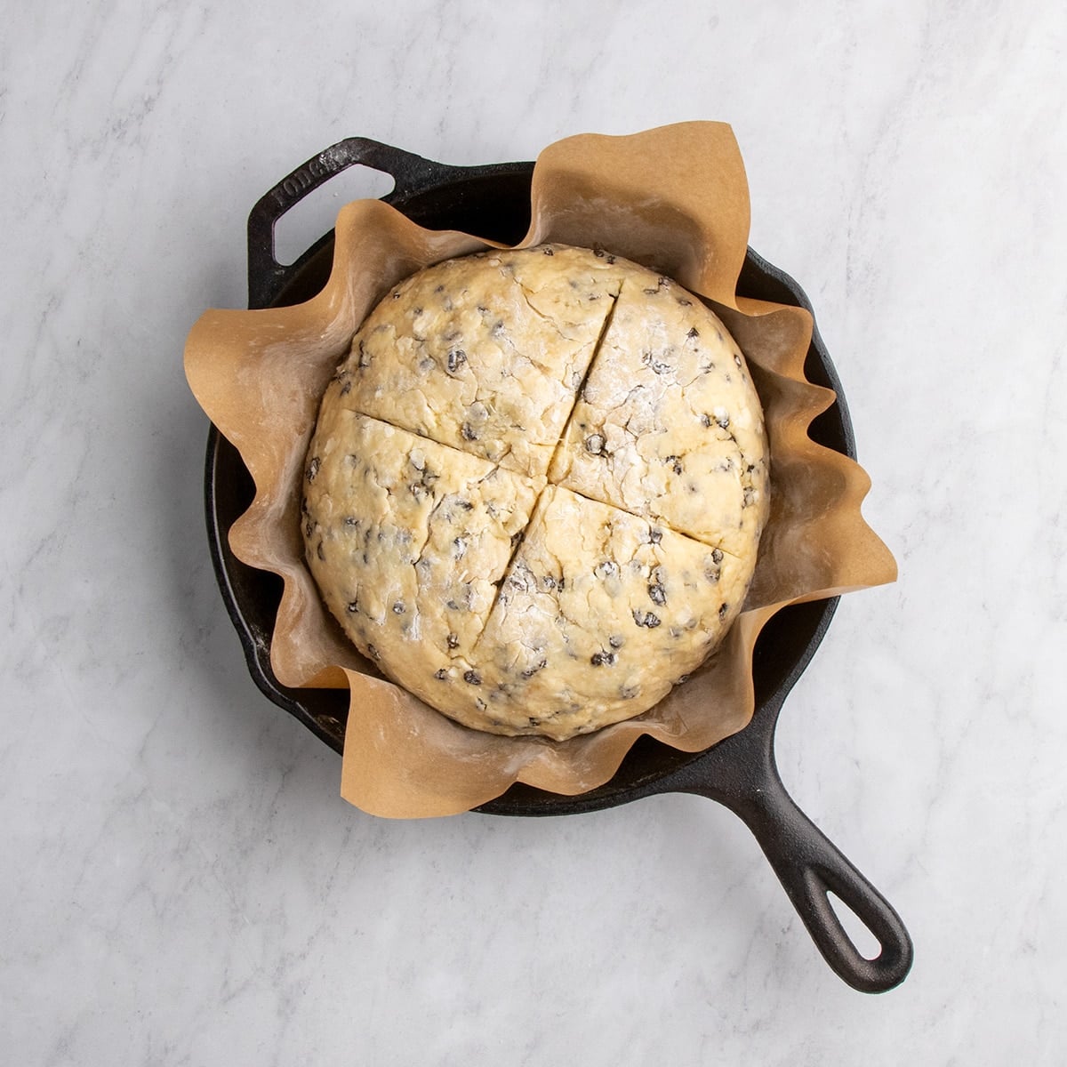 Round of dough with a cross cut into the top placed in a parchment-lined black round cast iron skillet.
