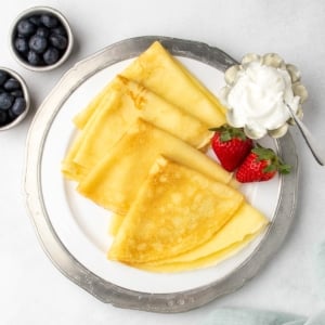 Overhead image of white plate with pewter rim, holding 4 folded gluten free crepes with strawberries and a small dish of white whipped cream, with bowls of blueberries nearby.