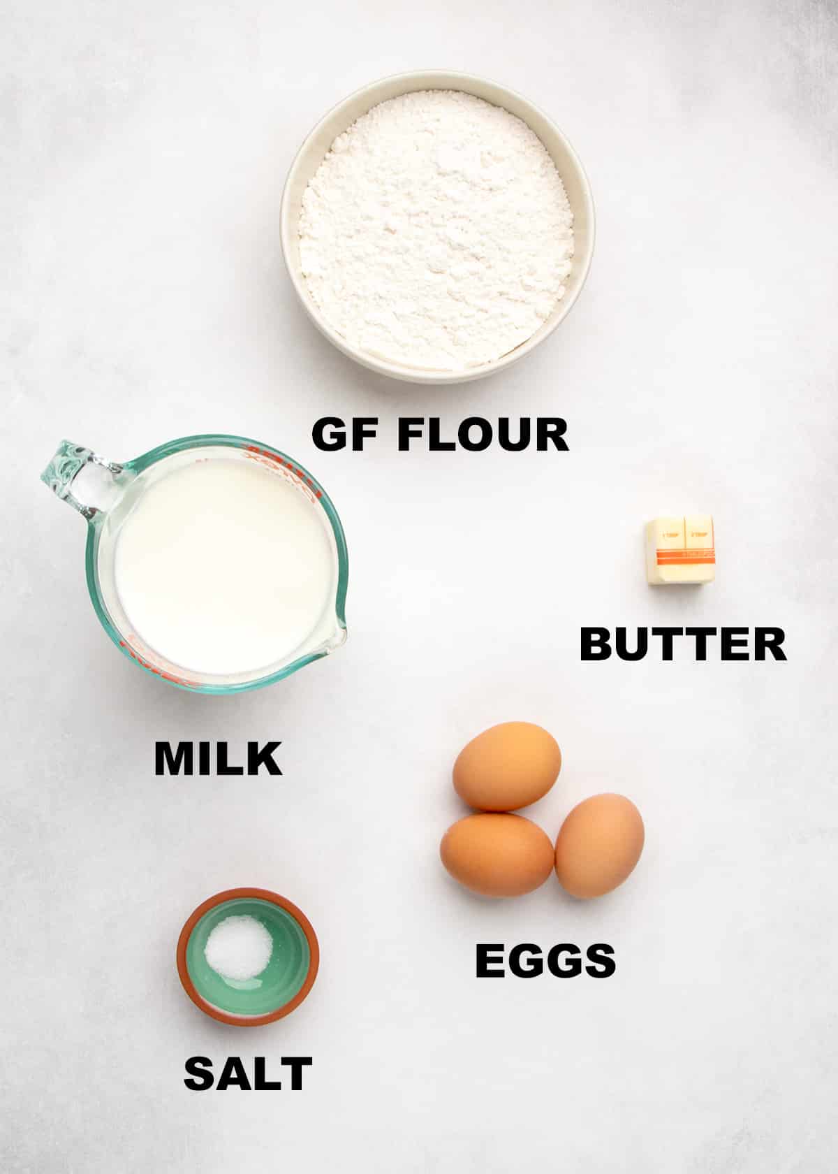 Measured ingredients including gluten free flour, milk, butter, eggs, and salt labeled on a light surface.