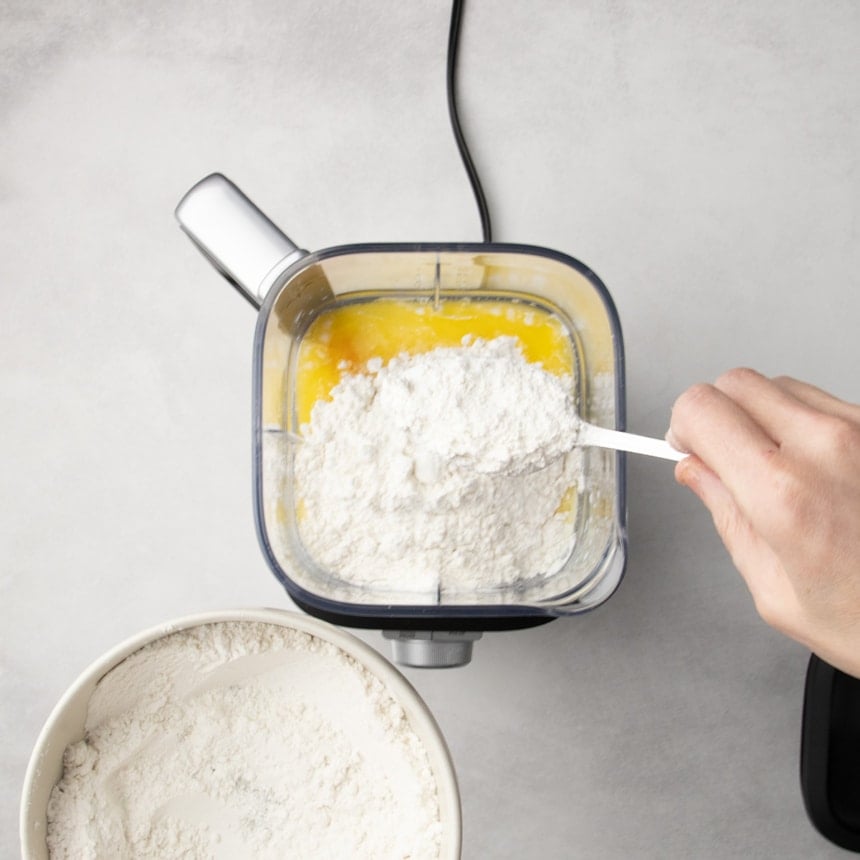 Hand holding a white scoop of flour over a blender container filled with yellow liquid, with a bowl of flour nearby.