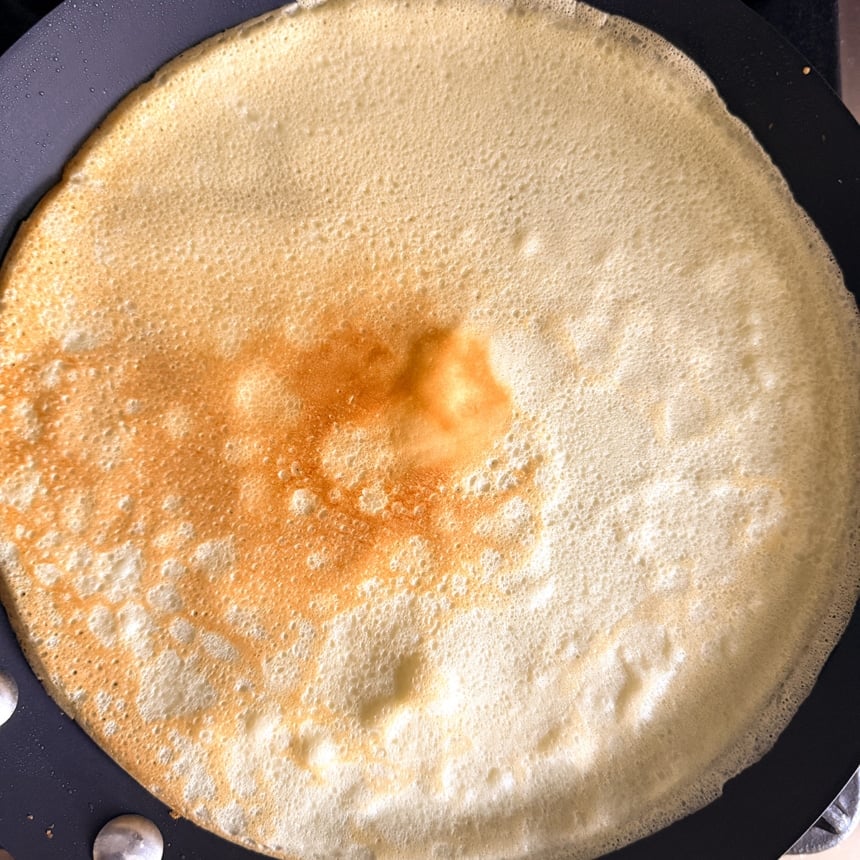 Crepe cooking in a skillet on the second side, with a lightly browned center and bubbles across the surface.