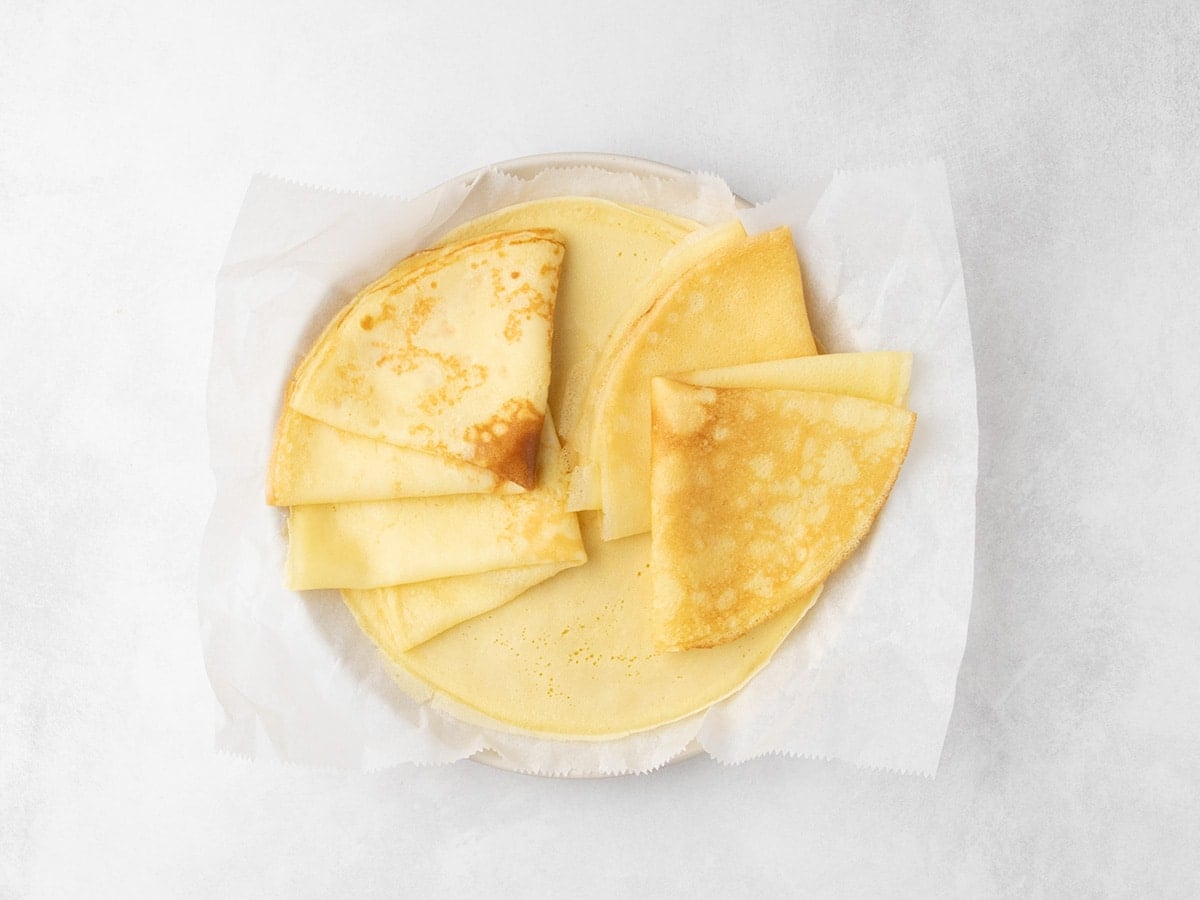 Four folded crepes on top of flat crepes sitting on parchment paper on a round plate.