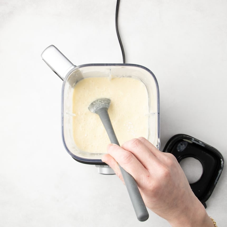 Hand holding a long gray spatula scraping down the inside of a blender container with smooth light yellow batter.
