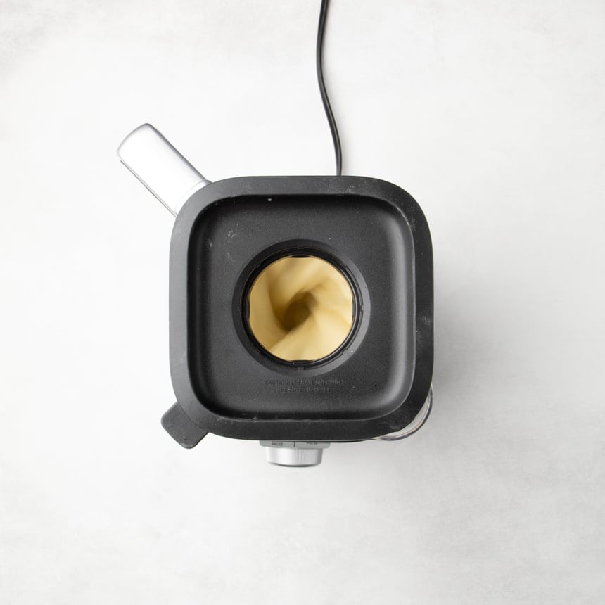 Blender with black lid on, showing light yellow batter blending again through the center opening.